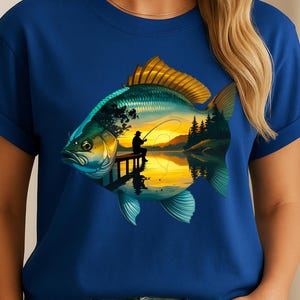 May include: Blue t-shirt featuring a detailed illustration of a fish with a scenic landscape inside. The fish design includes a fisherman on a dock, trees, and a sunset over a lake. The shirt is a solid blue color.
