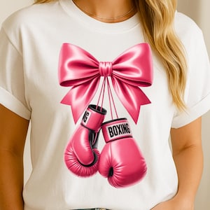 Puede incluir: Camiseta blanca con un gran lazo rosa y un par de guantes de boxeo rosas. Los guantes son negros y rosas con la palabra "BOXING" impresa en uno de ellos. La camiseta es de estilo casual.