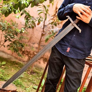 May include: A long, steel sword with a patterned blade and a cross-shaped hilt. The sword has a dark, aged appearance, suggesting it is a historical or replica weapon. The handle appears to be wrapped in a dark material, possibly leather or wood.