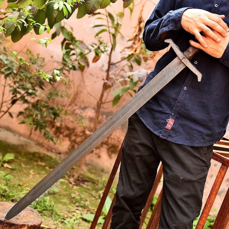 May include: A long, steel sword with a patterned blade and a cross-shaped hilt. The sword has a dark, aged appearance, suggesting it is a historical or replica weapon. The handle appears to be wrapped in a dark material, possibly leather or wood.