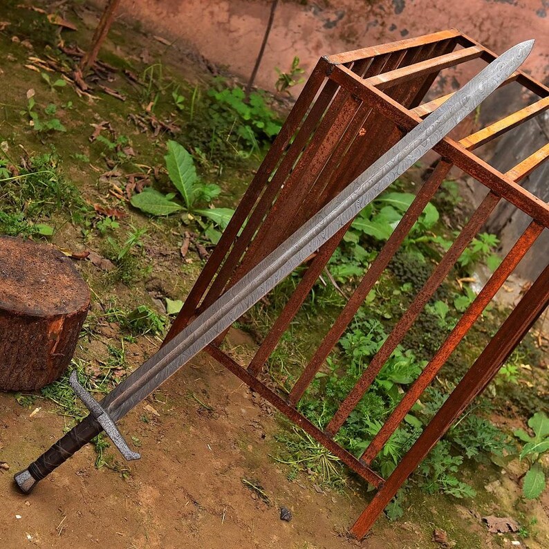 May include: A long, silver sword with a textured blade and a cross-shaped hilt. The handle is wrapped in dark brown leather. The sword is resting on a rusty metal structure and dirt.