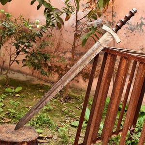 May include: A long, ornate sword with a patterned blade and a wooden handle. The sword is resting on a tree stump, with a rusty metal structure and greenery in the background. The blade has a unique, textured design.