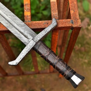 May include: Close-up of a medieval sword with a patterned steel blade and a crossguard. The handle is wrapped in dark brown leather. The sword is resting on a rusty metal structure.