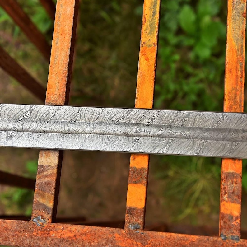 May include: Close-up of a patterned steel sword blade. The blade has a unique wood grain-like pattern. The sword is resting on a rusty metal surface with orange and brown bars.