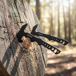May include: Two black hatchets embedded in a wooden target. The hatchets have a curved blade and a handle with cut-out designs. The text "DEER EDGE" is visible on the hatchets. The background is a blurred forest scene.