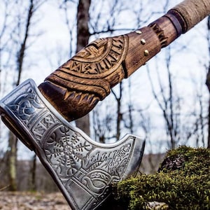 May include: A detailed close-up of a Viking axe. The axe head is silver with intricate engraved designs, while the wooden handle features carved patterns and is wrapped with a tan cord. The axe is resting on a bed of green moss.