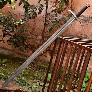 May include: A long, silver sword with a patterned blade and a dark brown handle. The sword features a crossguard and a pommel. The blade has a textured appearance. The sword is leaning against a rusty metal structure.
