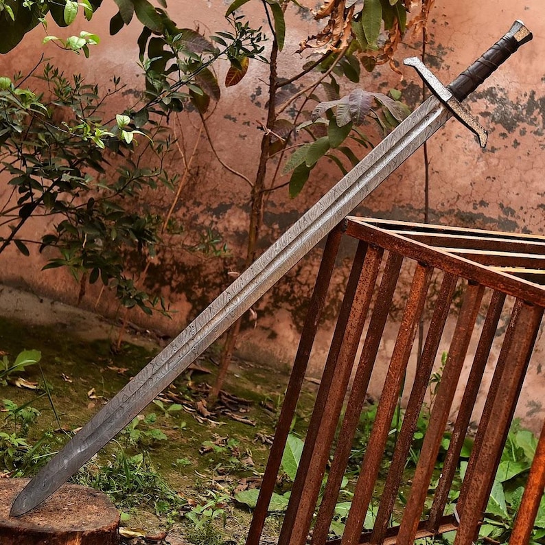 May include: A long, silver sword with a patterned blade and a dark brown handle. The sword features a crossguard and a pommel. The blade has a textured appearance. The sword is leaning against a rusty metal structure.