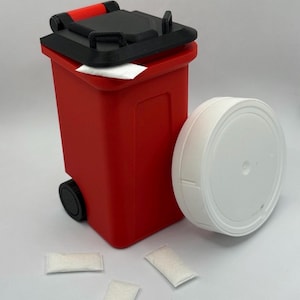 May include: A miniature red trash bin with a black lid and wheels, next to a white, round container. Three small white packets are scattered nearby. The bin has a red handle and a rectangular shape.