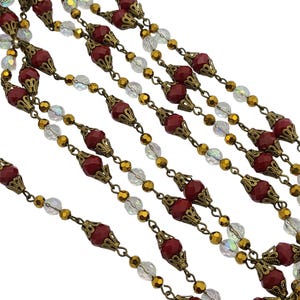 May include: Close-up of multiple strands of beaded jewelry. The strands feature alternating red, clear, and gold-colored beads. The red beads have a decorative, ornate metal cap. The beads are strung on a gold-colored chain.