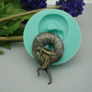 May include: A silicone mold for making a coiled dragon figurine. The mold is teal green and the dragon is a dark green, black, and gold color.
