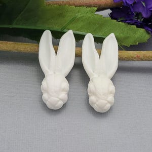 May include: Two white bunny head-shaped embellishments. Each bunny has long, upright ears and a detailed face. The bunnies are made of a smooth, solid material. The background is a light gray surface with a hint of green and purple.