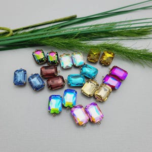 以下が含まれることがあります： 青、紫、金など、さまざまな色の長方形のイリデセントクリスタルラインストーンのコレクション。各石には、取り付け用の金属ループが付いています。背景には緑の葉が見えます。