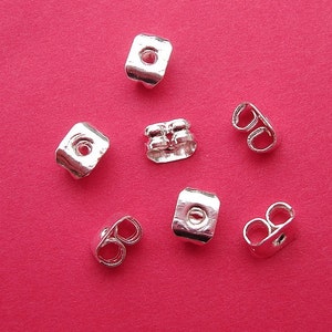 May include: A close-up of eight silver earring backs. The backs are designed to help keep earrings in place. Some are square shaped with a small hole in the center, while others are shaped like a figure eight.