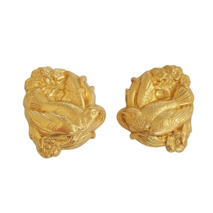 May include: Two gold-colored, bird-shaped decorative objects. Each object features a detailed relief of a bird surrounded by floral and leaf elements. The objects are likely earrings or decorative elements.