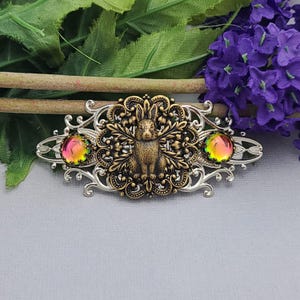 Könnte beinhalten: Eine silberfarbene Haarspange mit einem verzierten Filigranrahmen und einem zentralen, bronzefarbenen Hasenmotiv. Das Design umfasst zwei runde, schillernde Cabochons mit wechselnden rosa und grünen Farbtönen auf beiden Seiten des Hasen.