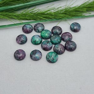 May include: A collection of round, polished gemstones in shades of purple, green, and gray. The stones are scattered on a white surface, with a few sprigs of green foliage in the background. These cabochons are suitable for jewelry making.