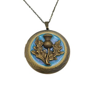 May include: A round antique bronze locket pendant on a matching chain. The locket features a raised bronze thistle emblem centered on a shimmering light blue enamel background. The outer edge of the locket has an intricate embossed floral pattern.