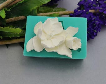 Può includere: Uno stampo in silicone rettangolare color verde acqua con un design floreale bianco in rilievo, raffigurante un grande fiore sbocciato con petali multipli e un bocciolo più piccolo sul lato per creare impronte dettagliate.