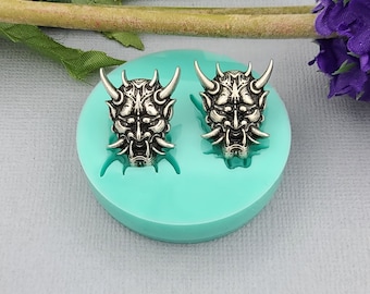 May include: A turquoise silicone mold with two impressions of a silver-colored oni mask. The oni mask has horns, fangs, and a fierce expression.