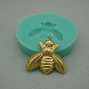 Puede incluir: Un abalorio de abeja de color dorado con una superficie texturizada. La abeja tiene un cuerpo redondo con alas y un pequeño bucle en la parte superior para colgar.