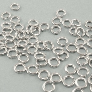 May include: A large quantity of silver jump rings, scattered on a light gray surface. The jump rings are small and circular, with a gap in the circle.