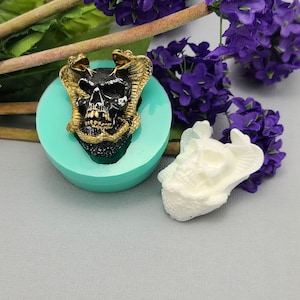 May include: A silicone mold for making a skull with snakes around it. The mold is turquoise and the skull is black with gold accents. A white resin skull is also shown.