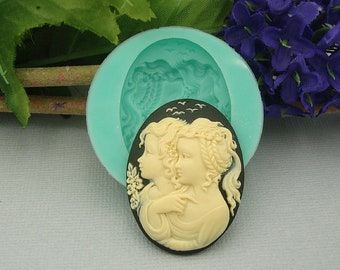 May include: A cameo mold and a finished cameo. The mold is a light teal color, circular, and features a relief of two women. The finished cameo is oval, with a black background and a cream-colored relief of two women with floral and bird details.