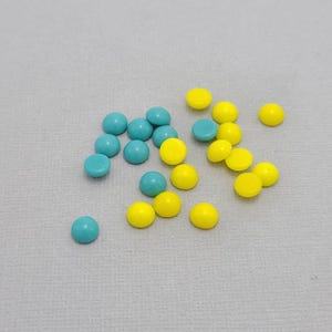 Puede incluir: Una colección de pequeños caramelos redondos turquesa y amarillos esparcidos sobre una superficie gris claro. Los caramelos están espaciados uniformemente y parecen estar hechos de un material liso y brillante. La imagen está bien iluminada, sin sombras.