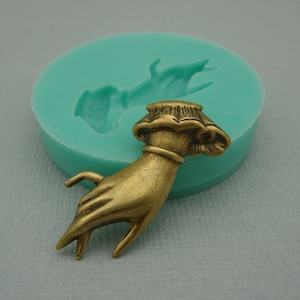 May include: A gold-toned metal hand with a decorative cuff and a raised design on the back of the hand. The hand is in a reaching gesture with the fingers extended.