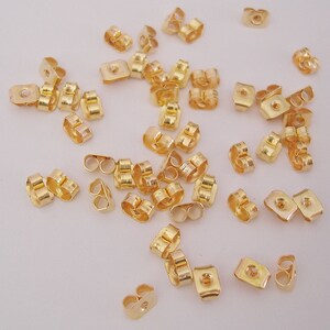 May include: A large quantity of gold-colored earring backs. The backs are made of metal and have a small loop on each side. They are designed to be used with post earrings.