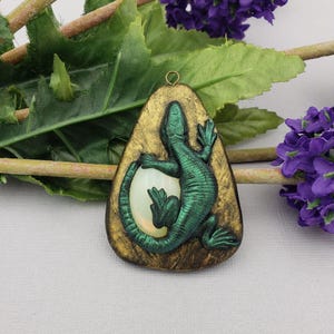 May include: A handcrafted pendant featuring a green lizard design over a white oval, set against a gold-colored background. The pendant has a teardrop shape and is suspended from a small gold loop. The lizard is detailed with textured scales.
