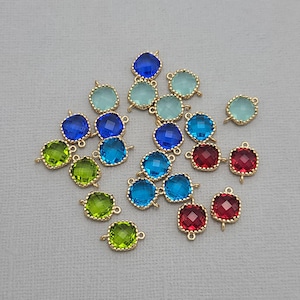 以下が含まれることがあります： 金色の金属フレームにカラフルな正方形の宝石チャームのコレクション。宝石は青、緑、赤の色合いです。各チャームには、ジュエリーに取り付けるための小さなループが付いています。