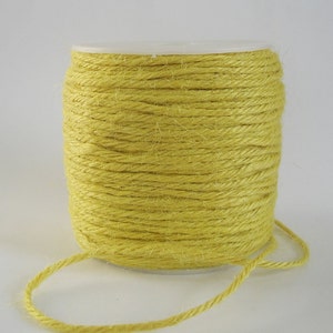 May include: A spool of yellow twine, tightly wound around a white plastic core. The twine is a muted yellow color, and a length of the twine extends from the spool. This craft supply is suitable for various projects.