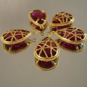 以下が含まれることがあります： 幾何学模様と濃いピンク色のクリスタルをあしらった、ゴールドトーンの涙型のペンダントが5つ。ペンダントの上部には、チェーンやコードに取り付けるためのループが付いています。