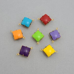 May include: Seven colorful square gemstone charms in turquoise, red, orange, green, purple, and yellow. Each charm has a gold-colored metal frame with loops for attaching to jewelry. The charms are arranged on a light gray surface.