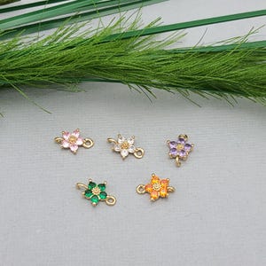 以下が含まれることがあります： カラフルな宝石の花びらを持つ、5つの小さな金色の花のチャーム。ピンク、白、紫、緑、オレンジの花びらが特徴です。各チャームには、ジュエリーやその他の工芸品に取り付けるための小さなループが付いています。