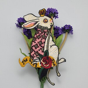 May include: White embroidered patch of a rabbit wearing a pink and white dress with a crown on its head. The rabbit is holding a red rose and a banner that says "The True".