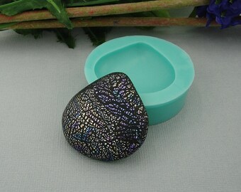 Könnte beinhalten: Eine türkisfarbene Silikonform zum Herstellen von Schmuck aus Harz. Die Form ist tropfenförmig und hat ein detailliertes Design. Ein schwarzes Harzstück mit einem silbernen und schillernden Muster liegt neben der Form.