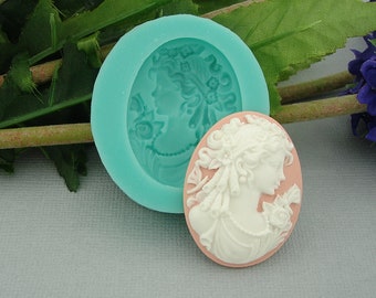 May include: A light blue silicone mold and a finished cameo. The mold is round with a detailed profile of a woman with flowers and ribbons. The cameo is oval, with a white profile on a pink background, and a floral design.