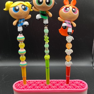 May include: Three pens featuring cartoon character figures, each with a unique design. The pens are in a pink pen holder. The first pen has a yellow figure, the second has a green figure, and the third has an orange figure. Each pen has a different colored shaft and decorative beads.