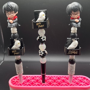 May include: Three decorative pens with Harry Potter-themed designs. Each pen features a figurine of Harry Potter, a white owl, and the words "Harry Potter" or "Always". The pens have black, white, and silver accents, and are displayed in a pink pen holder.