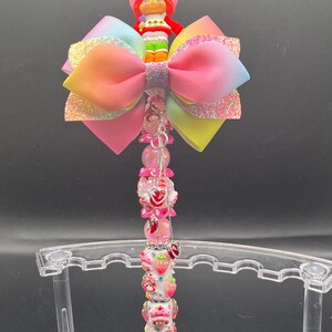 May include: A decorative pen featuring a Strawberry Shortcake character figurine atop a large, multi-layered bow in rainbow colors. The pen's shaft is adorned with pink and red beads, strawberries, and heart-shaped charms. The pen is displayed on a clear stand.