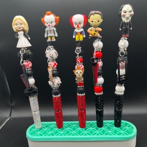Op de afbeelding: Vijf decoratieve pennen met figuren van horrorfilmkarakters. Elke pen heeft een ander karakterfiguur, een kralenontwerp en een met strass bedekte schacht. De pennen worden gepresenteerd in een groen-witte pennenhouder.
