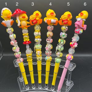 May include: A collection of six decorative pens with yellow barrels and various toppers. The toppers include a chick hatching from an egg, a mushroom, a chick with fries, and a chick on a scooter. The pens have colorful beaded accents.