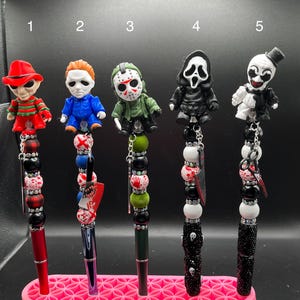 Op de afbeelding: Vijf decoratieve pennen met figuren van horrorfilmkarakters. Elke pen heeft een ander karakterfiguur, een kralenontwerp en een penpunt. De pennen worden tentoongesteld in een roze pennenhouder.