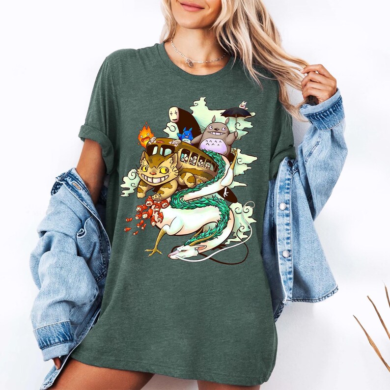 K&ouml;nnte beinhalten: Dunkelgr&uuml;nes T-Shirt mit einer farbenfrohen Grafik von animierten Charakteren. Das Design umfasst eine gelbe katzenartige Kreatur, einen Bus und einen wei&szlig;en Drachen. Das Shirt ist ein bequemer, l&auml;ssiger Stil.