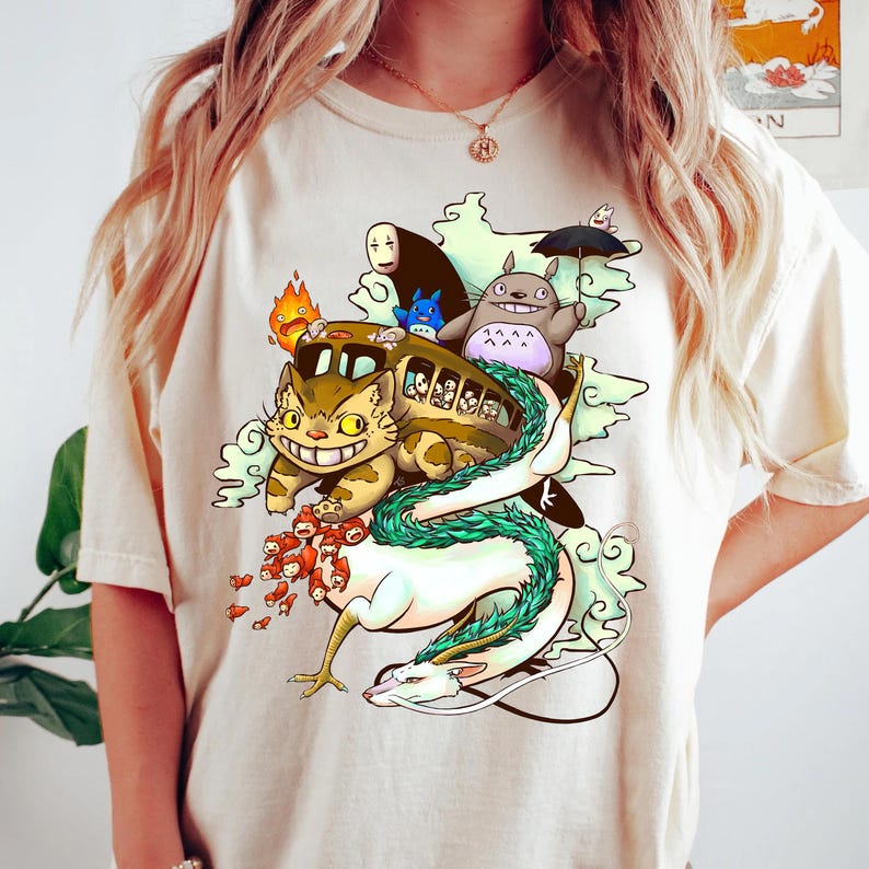 K&ouml;nnte beinhalten: Cremefarbenes T-Shirt mit einer farbenfrohen Grafik von Charakteren aus einem japanischen Animationsfilm. Das Design umfasst einen Katzenbus, Totoro, einen Drachen und andere skurrile Kreaturen. Das Kunstwerk ist detailliert und lebendig.