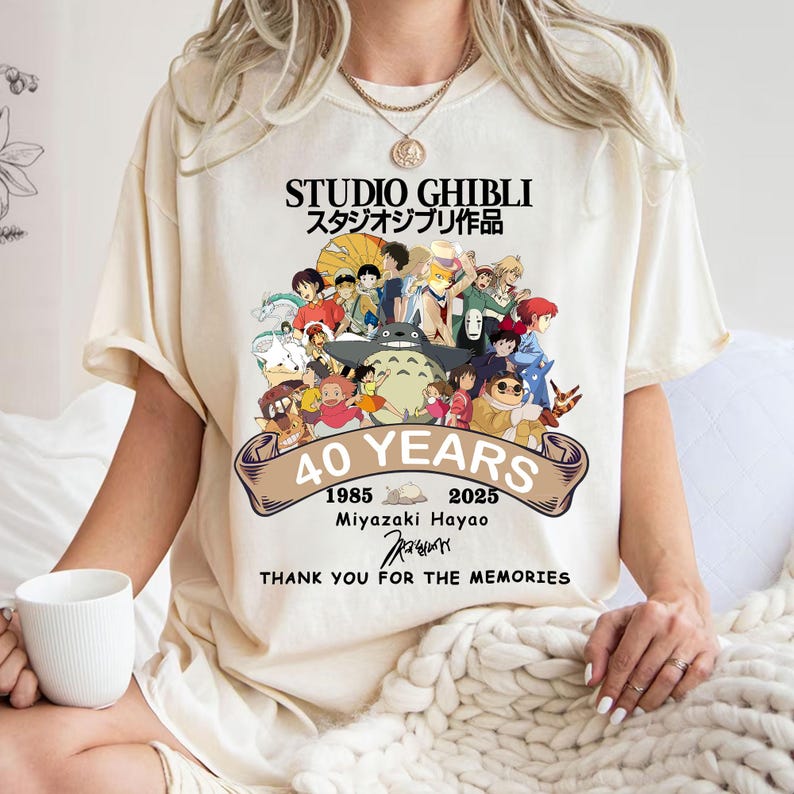 May include: Cream-colored t-shirt featuring a colorful graphic celebrating Studio Ghibli's 40th anniversary. The design includes characters from various Ghibli films, the text "STUDIO GHIBLI", and the years 1985-2025. The shirt also includes the text "THANK YOU FOR THE MEMORIES".