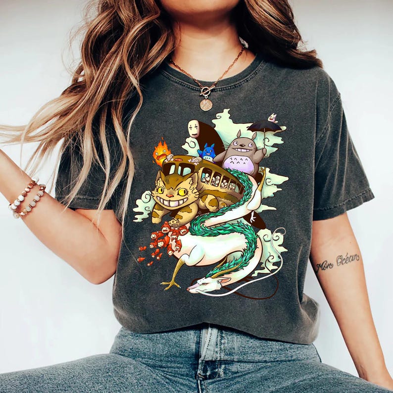 K&ouml;nnte beinhalten: Dunkelgraues T-Shirt mit einem farbenfrohen Grafikdesign von Charakteren aus einem beliebten Zeichentrickfilm. Das Design umfasst einen katzenf&ouml;rmigen Bus, einen Drachen und andere skurrile Kreaturen. Das Shirt ist aus weichem Material.